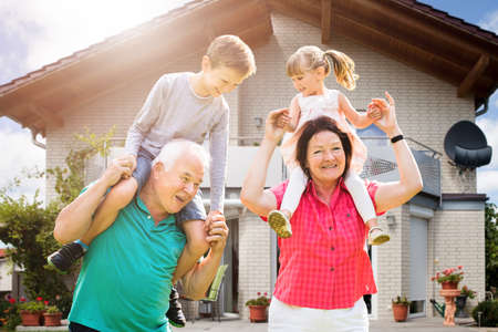 Smiling Grandchildren's Sitting On Grandparent's Shoulder Enjoying Outside Their Houseの写真素材