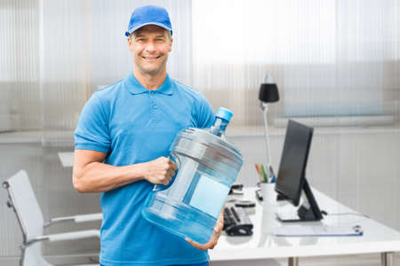 Portrait Of A Smiling Mature Delivery Man Holding Large Water Bottle In Officeの写真素材
