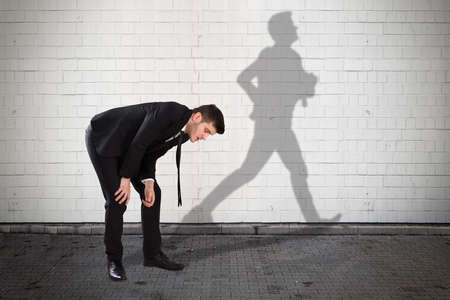 Young Tired Businessman With Shadow Of Man Running Formed On Wallの写真素材
