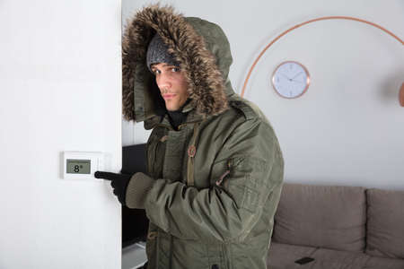 Young Man In Warm Clothing Pointing To Current Room Temperature In Digital Thermostat At Home