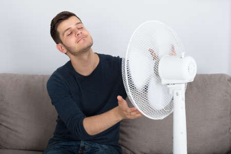 Young Man Sitting On Couch Enjoying Breeze With Electric Fan At Homeの写真素材
