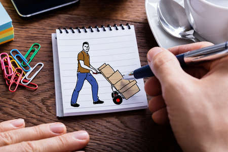 Close-up Of A Person's Hand Drawing Delivery Man Moving Trolley Full Of Boxes On Notepad