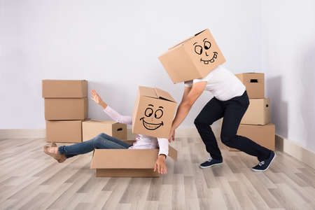 Man Wearing Cardboard Box Pushing A Woman Sitting On Cardboard Box At Homeの写真素材