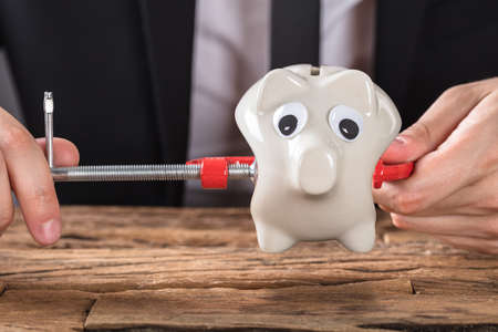 Close-up Of Businessperson Squeezing Piggy Bank In Clamp Tool At Deskの写真素材