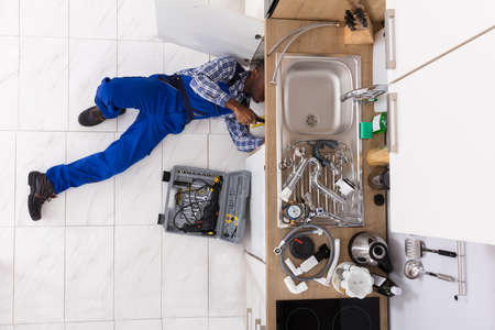 High Angle View Of Handyman Lying On Floor Repairing Sink In Kitchen With Toolboxの写真素材