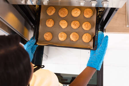 Human Hand Wearing Blue Gloves Taking Out Tray Of Baked Cookies From Microwave Ovenの写真素材