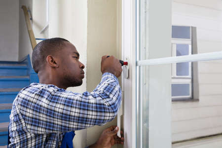 Young African Handyman In Uniform Fixing Glass Window With Screwdriver