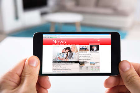 Close-up Of A Person Holding Mobile Phone With Online News On Screenの写真素材