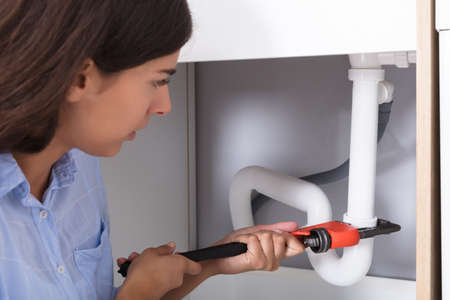 Close-up Of Young Woman Tightening Sink Pipe With Monkey Wrench In Kitchenの写真素材