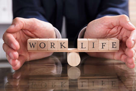 Close-up Of A Businessperson's Hand Protecting Balance Between Life And Work On Seesaw Over Desk