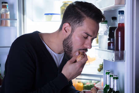 Close-up Of A Young Man Eating Cookie Near Open Refrigerator