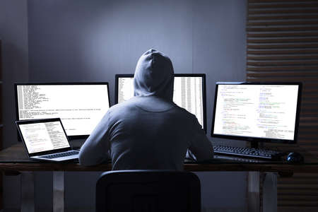 Rear View Of A Hacker Using Multiple Computers For Stealing Data On Desk