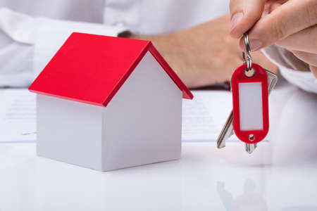 Human Hand Holding House Keys With House Model On Desk