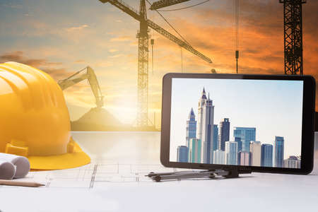 Close-up Of Digital Tablet And Hardhat On Blueprint In Front Of Construction Site