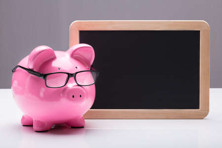 Close-up Of A Piggybank With Eyeglasses Near Blank Slate On Deskの写真素材
