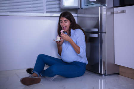 Young Woman Sitting On Floor Eating Slice Of Cake In Kitchen