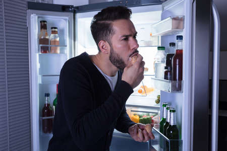 Close-up Of A Young Man Eating Cookie Near Open Refrigeratorの写真素材