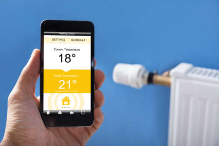 Close-up Of Person Hand Setting Temperature Of Thermostat Using Mobilephone At Homeの写真素材