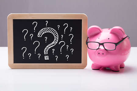Close-up Of Piggy Bank With Eyeglass And Blackboard With Question Marks On Deskの写真素材
