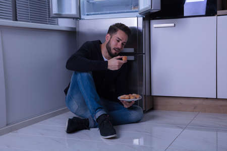 Young Man Sitting On Floor Eating Cookie In Kitchenの写真素材