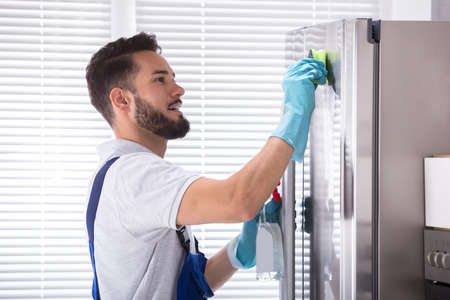 Side View Of A Young Male Janitor Cleaning Refrigerator In Kitchenの写真素材