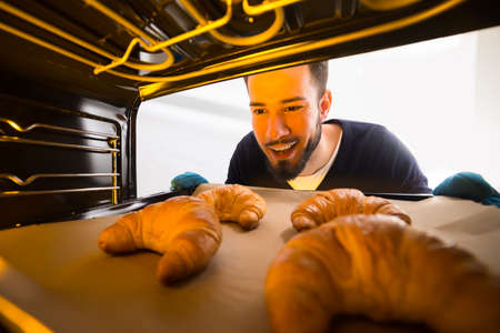 Happy Young Man Taking Out Tray Of Baked Croissants From Ovenの写真素材