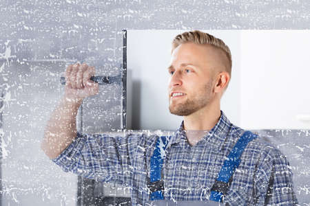 Smiling Young Worker Cleaning Glass Window With Squeegee In Kitchen