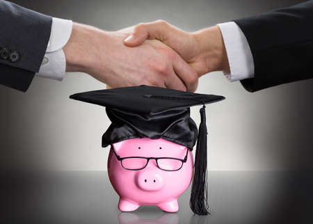 Piggy Bank With Black Graduation Hat And Eyeglasses In Front Of Two People Shaking Handsの写真素材