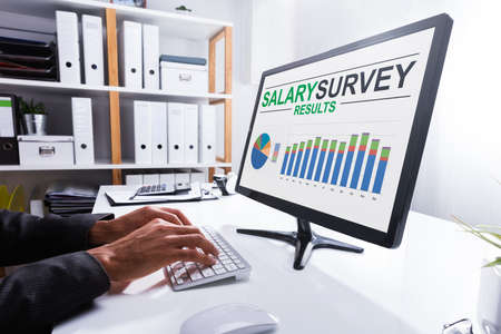 Close-up Of A Businessperson's Hand Analyzing Salary Survey Results On Computer At Workplace