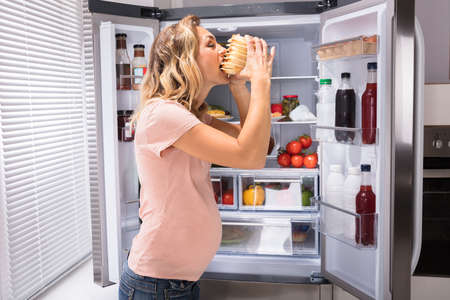 Pregnant Woman Eating Sandwich In Front Of Open Refrigeratorの写真素材