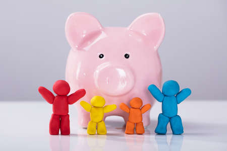 Row Of Colorful Clay Family In Front Of Pink Plastic Piggybank Over The White Deskの写真素材