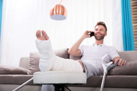 Happy Young Man With Broken Leg Sitting On Sofa Talking On Smartphoneの写真素材