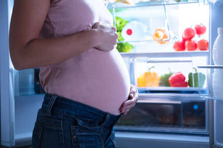Close-up Of Pregnant Woman Standing Near Refrigerator Full Of Healthy Foodの写真素材
