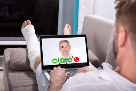 Close-up Of A Young Man Lying On Sofa Video Conferencing With Doctor On Laptop