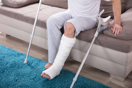 Young Man With Broken Leg Using Crutches To Get Up From Sofa