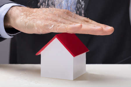 Close-up Of Male's Hand Protecting The House Model On Desk From Falling Waterの写真素材