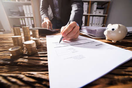 Close-up Of A Businessperson's Hand Signing Document With Stacked Golden Coins On Wooden Deskの写真素材