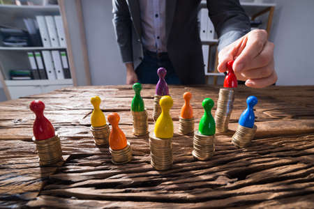 Close-up Of A Businessperson's Hand Placing Red Figurine Pawn On Top Of Stacked Golden Coinsの写真素材