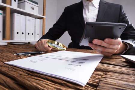 Businessperson's Hand Analyzing Bill With Magnifying Glass On Wooden Deskの写真素材