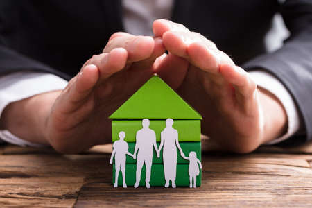Businessperson's Hand Protecting House Model And Family Paper Cut Out On Wooden Deskの写真素材