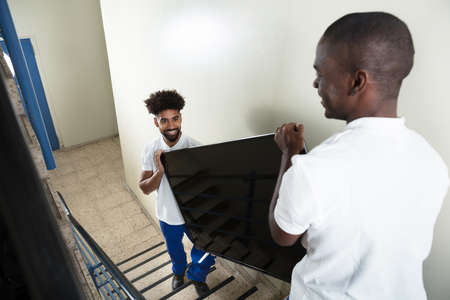 Close-up Of Two Male Young Movers Carrying Television While Climbing Steps At Homeの写真素材