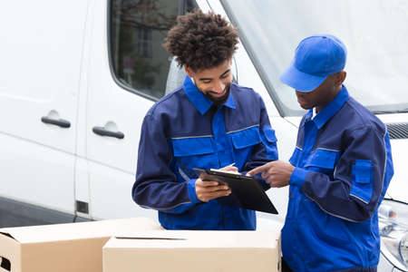 Delivery Man Writing On The Clipboard Over The Cardboard Boxes Standing By The Truckの写真素材