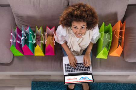 Happy Young Woman Using Laptop With Multi Colored Shopping Bags On Carpetの写真素材