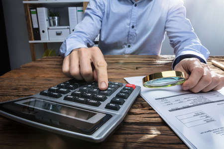 Close-up Of A Businessperson's Hand Holding Magnifying Glass While Using Calculator At Workplaceの写真素材