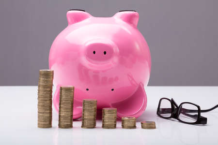 Upside Down Piggy Bank On Table With Eye Glasses And Stacked Coins On Tableの写真素材