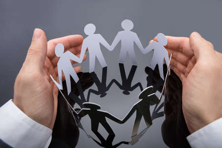 High Angle View Of A Businessperson's Hand Protecting Cut-out Figures On Deskの写真素材