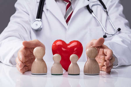 Doctor's Hand Protecting Wooden Family And Red Heart On White Deskの写真素材