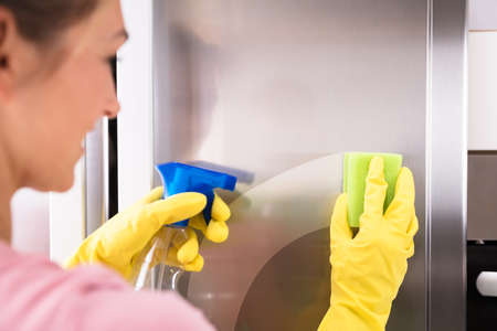 Close-up Of Woman's Hand Wearing Yellow Gloves Cleaning Stainless Steel Refrigerator With Spongeの写真素材