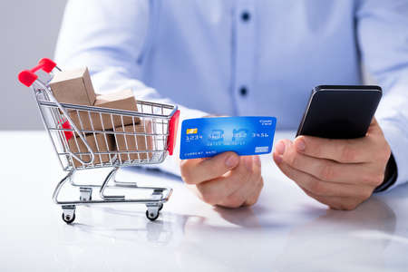 Hand Holding A Credit Card And Cell Phone For Online Shopping With Miniature Shopping Trolleyの写真素材