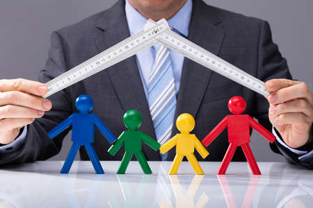 Businessperson's Hand Holding Measurement Tape Over Multi Colored Human Figures On White Deskの写真素材